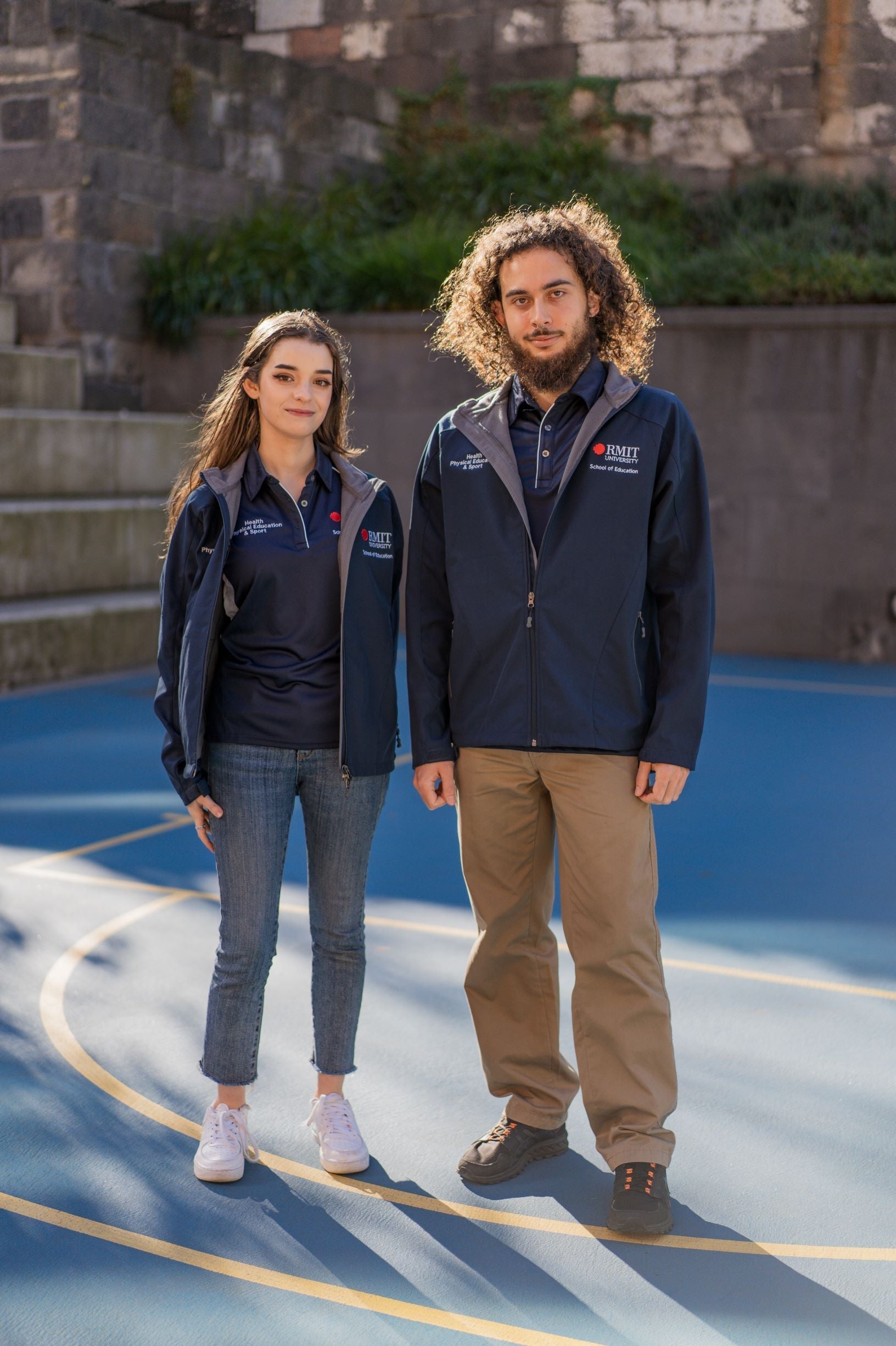 Full front view of a male and a female model wearing the Health, Physical Education & Sport Uniform – Jacket, both standing on campus and looking into the camera. The female model has her head slightly turned to the left, while the male model faces slightly right. The navy jackets feature a white embroidered “Health, Physical Education & Sport” on the right chest and the RMIT University logo on the left chest, worn over matching polos.